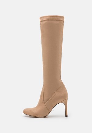 Botte beige en daim montant jusqu'au genou, avec un bout pointu, un talon aiguille fin et un design sans coutures. Présente une texture lisse et aucun élément métallique visible.