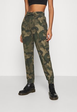 Person iført grønne camouflage joggerbukser, sorte snørestøvler og en sort cropped top, stående i et neutralt indendørs miljø.