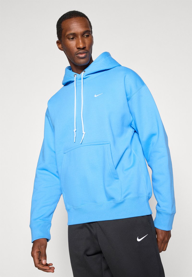 Blaue Baumwollkapuzenjacke mit einer Fronttasche, weißen Kordeln und einem kleinen weißen Logo auf der Brust. Kombiniert mit schwarzen Hosen.
