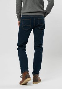 Jeans en denim bleu avec une coupe slim, présentant des poches arrière avec des coutures en angle. Portés avec des bottes à lacets marron et un pull gris.