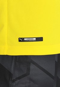 Ljusgul sporttröja med en strukturerad yta. Har en svart etikett med en logotyp och produktnummer längst ner, mot mörkmönstrade shorts.