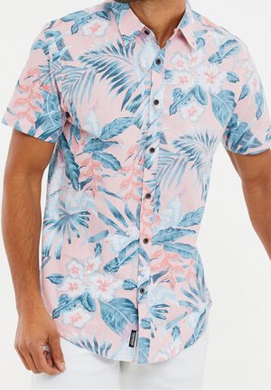 Homme portant une chemise rose à manches courtes avec imprimé floral tropical bleu et blanc.
