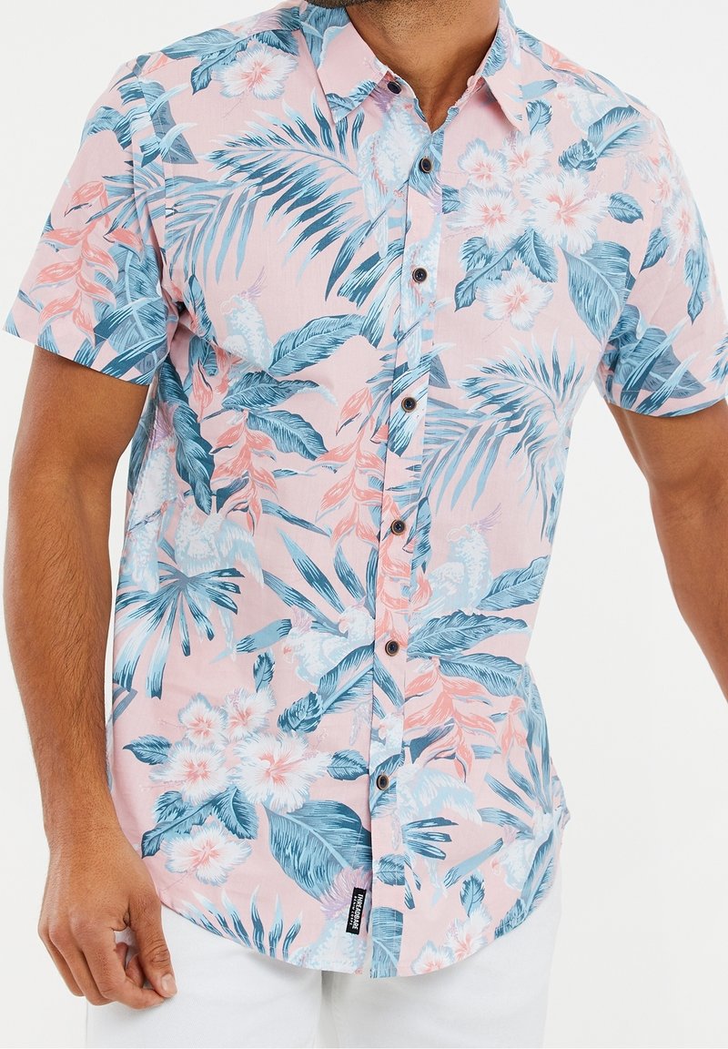 Homme portant une chemise rose à manches courtes avec imprimé floral tropical bleu et blanc.