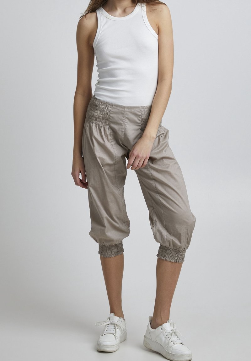 Jeune personne portant un haut blanc sans manches, un pantalon beige court avec des poignets élastiques et des baskets blanches, debout devant un fond uni.