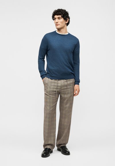 Άνδρας που φοράει μπλε πουλόβερ με μακριά μανίκια, μπεζ καρό παντελόνι και μαύρα παπούτσια, στέκεται με τα χέρια στις τσέπες κοιτάζοντας πλάγια.