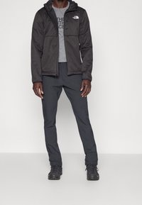 Giacca nera con felpa grigia con zip, t-shirt grigia e pantaloni scuri. Presenta un logo sul petto e scarpe nere robuste, adatte ad attività all'aperto.