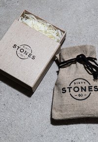 Boîte en carton avec une finition texturée brun clair, contenant de la paille blanche, à côté d'un sachet en tissu beige avec un cordon noir et un logo imprimé.