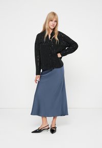 Camisa preta de mangas longas com recortes verticais, combinada com uma saia midi azul em cetim. Modelo usa saltos slingback pretos. Fundo simples.