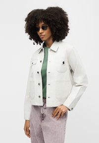 Γυναίκα με στρογγυλά μαύρα γυαλιά ηλίου, ελαφρύ ριγέ σακάκι με κουμπιά πάνω από πράσινο πουκάμισο και ελαφριά μωβ παντελόνια ψηλής μέσης, που στέκεται μπροστά σε λευκό φόντο.