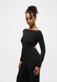 Γυναίκα με σγουρά μαλλιά σε ψηλό σινιόν, ντυμένη με εφαρμοστό μακρυμάνικο μαύρο τοπ και παντελόνι ψηλόμεσο, ευρύ, σε λευκό φόντο.