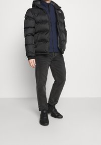 Svart pufferjacka med huva, med dragkedja fram, elastiska muddarna och logotypdetalj. Bärs över en marinblå tröja och matchas med svarta jeans.