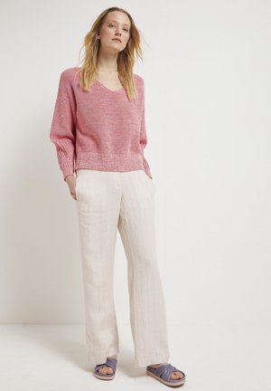 Rosa Strickpullover mit V-Ausschnitt und lockerem Schnitt, kombiniert mit off-white gestreiften Hosen und lila Rutschsandalen. Strukturiertes Stoffdetail sichtbar.