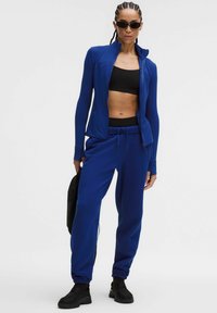 Veste de sport bleu roi à col montant, assortie d'un pantalon de survêtement. Crop top noir et chaussures complètent la tenue sportive.