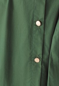 Zelená látka s hladkým povrchem, zdobená světle barevnými kulatými knoflíky jako kovovými prvky na přední straně. Jednoduchý, čistý design.