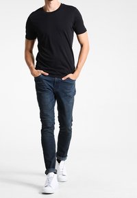 Muž v černém tričku s krátkým rukávem, tmavě modrých džínách s manžetami a bílých teniskách, kráčející s rukama v kapsách na bílém pozadí.
