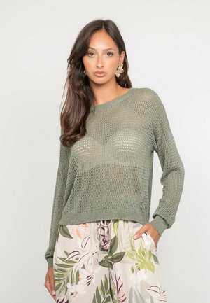 Femme aux longs cheveux noirs portant un pull vert en maille ajourée, un pantalon large à motifs floraux et de grandes boucles d'oreilles en forme de feuille, debout avec une main dans la poche.