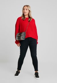 Red knitted oversized sweater, black skinny jeans, and black sneakers. The patterned bag features a chain strap and contrasting colors.