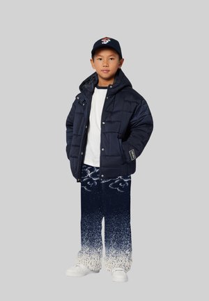 Marineblauwe pufferjas, wit T-shirt, geblokte blauw-witte broek en witte sneakers. Een pet met grafisch ontwerp maakt de outfit compleet.