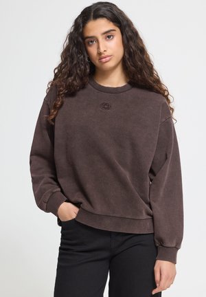 Ung kvinde med langt krøllet hår iført en løs brun sweatshirt og sorte bukser, står med den ene hånd i lommen.