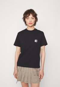 T-shirt en coton noir avec un col rond, des manches courtes et une poche frontale avec un patch logo. Porté avec une jupe beige plissée.