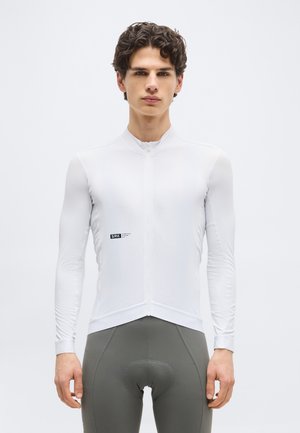 Junger Mann mit lockigem Haar, gekleidet in ein eng anliegendes weißes Langarm-Radtrikot und graue gepolsterte Radhosen, stehend vor einem schlichten hellen Hintergrund.