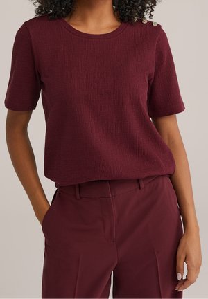 Kobieta ubrana w bordowy, teksturowany top z krótkim rękawem i złotymi guzikami na ramieniu, w połączeniu z bordowymi spodniami z wysokim stanem.