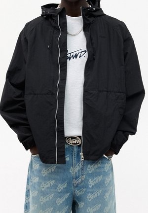 Person trägt schwarze Jacke mit Reißverschluss über einem weißen T-Shirt mit Grafik, eine Kettenkette und blaue Jeans mit weißem Aufdruck sowie einen Gürtel mit Logoschnalle.