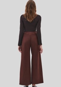 Vrouw met lang bruin haar, gekleed in een aansluitend zwart langslavend bovenstuk en brede bruine broek, staande tegen een lichtgrijze achtergrond.