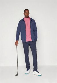 Námornícka bunda na zips, svetloružové polokošeľa, námornícke nohavice a biele golfové topánky. Muž drží golfovú palicu, stojí pred jednoduchým pozadím.