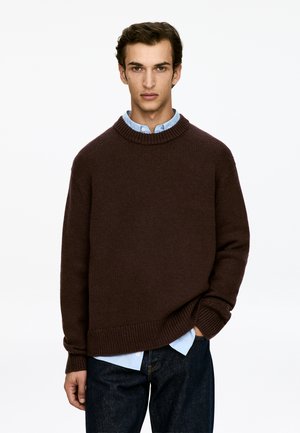 Brauner Strickpullover mit einem gerippten Rundhalsausschnitt, getragen über einem hellblauen Hemd mit Kragen. Dunkelblaue Jeans aus Denim mit leichter Struktur und lässiger Passform.