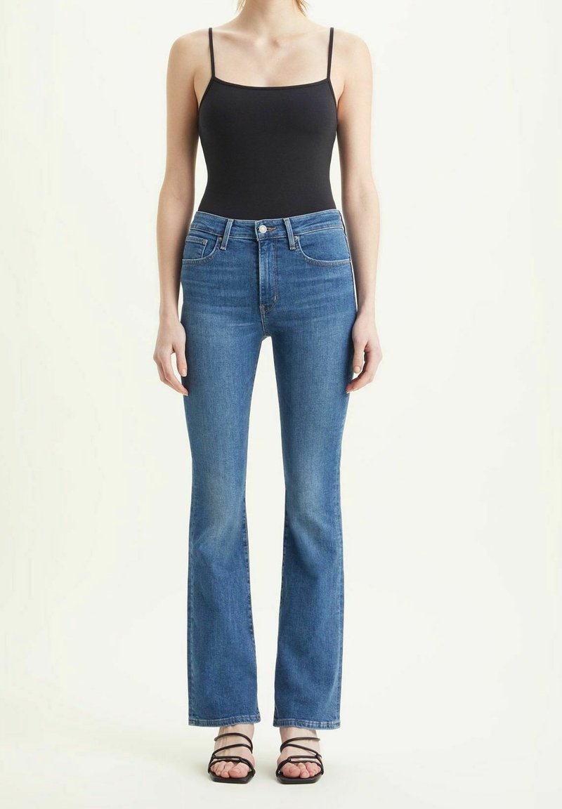 Pantalones vaqueros acampanados azules con cintura de tiro medio, cuatro bolsillos y textura de mezclilla de lavado claro, combinados con una camiseta negra ajustada y sandalias de tiras.