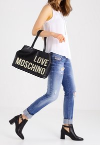 Černá kožená taška přes rameno s nápisem "LOVE MOSCHINO" zlatou barvou. Nosená s světlými opranými džíny a kotníkovými botami s blokovým podpatkem.