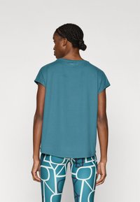 Τιρκουάζ μπλούζα με κοντά μανίκια και στρογγυλή λαιμόκοψη, φορεμένη με κολάν με γεωμετρικά σχέδια σε μπλε και λευκό.