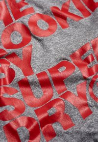 Szary materiał o gładkiej teksturze z wyraźnym, błyszczącym czerwonym napisem "SUPER DRY" oraz "LOOKING" w dużej czcionce.
