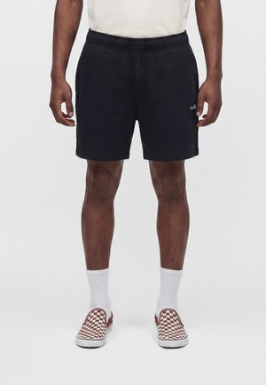 Male wearing black shorts, white socks, and red-and-white checkered slip-on shoes, standing with hands relaxed by sides.