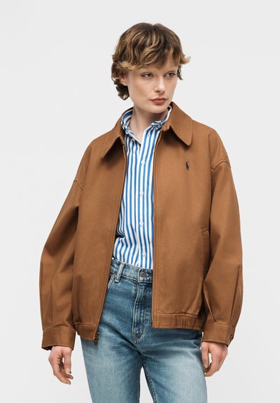 Άτομο που φοράει καφέ μπουφάν με φερμουάρ πάνω από μπλε και λευκό ριγέ πουκάμισο με κουμπιά και ανοιχτόχρωμα μπλε τζιν, στέκεται μπροστά σε απλό φόντο.