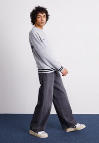 Grå trøje med sorte detaljer, løse sorte jeans og grå og gule sneakers. Modellen står på et blåt tæppe med en simpel baggrund.