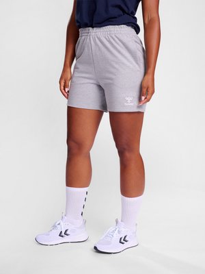 Personne portant un short gris, des chaussettes de sport blanches et des baskets blanches, debout devant un fond uni.