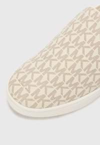Baskets slip-on avec un motif géométrique beige et crème, présentant une texture lisse et une semelle en caoutchouc blanc, marquées "Michael Kors."