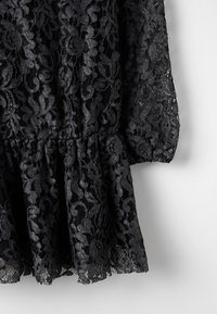 Svart blommig spets tyg med samlad midja och puffärmsdetalj på en ljusgrå bakgrund.