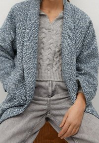 Cardigan a spina di pesce blu e bianco strutturato con spalle cadenti, indossato sopra un pullover grigio chiaro a treccia e jeans grigi a vita alta.