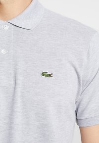 Sivá polo košeľa s textúrovanou tkaninou, s dvojitým gombíkom a ikonickým zeleným logo krokodíla na ľavej prednej strane.