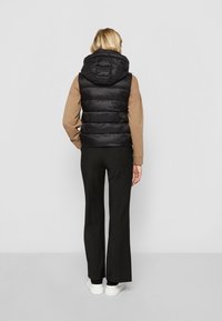 Gilet nero imbottito con colletto alto e cappuccio incorporato, indossato sopra un top beige e pantaloni neri. Texture liscia e design trapuntato visibile.