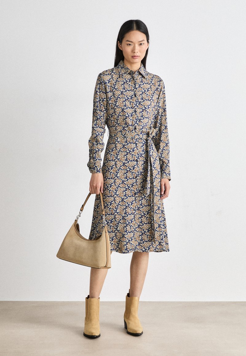 Rochie cu model floral în crem, albastru și auriu, cu guler, mâneci lungi, talie legată și tiv până la genunchi. Ținând o geantă de umăr bej.