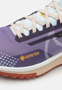 Fioletowy but biegowy z siateczkową cholewką, niebieskimi akcentami, beżową podeszwą pośrednią i pomarańczowym logo GORE-TEX. Posiada teksturowany fioletowy wzór i widoczne sznurowadła.