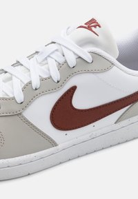 Nike sneaker met een witte leren bovenkant met grijze accenten, een bruine suède swoosh, witte veters en een gestructureerde rubberen zool.