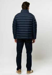 Mees näoga eemale, seljas tumesinine pufferjakk, tumedad teksad ja beežid spordijalatsid, helel taustal.