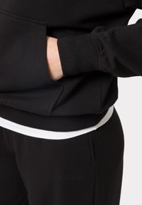 Černá mikina se klokaní kapsou, žebrovanými manžetami a pasem. Obsahuje decentní embosované textové logo značky. Vrstvená s bílým tričkem pod ní.