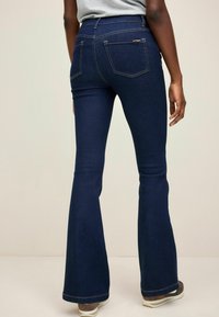 Jeans a zampa d'elefante blu scuro realizzati in denim. Presentano una vita alta, due tasche posteriori e cuciture a contrasto. Indossati con sneakers chiare.