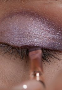 Close-up van een oog met glanzende paarse oogschaduw. Een kwast is boven de wimpers gepositioneerd, wat fijne details en textuur benadrukt.
